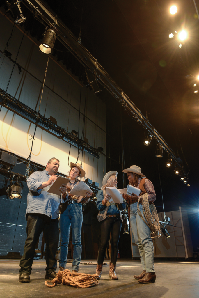 Professor Richard Perez stands on a stage without a set, with cast members Sam Hill ’18, Olivia Lehnertz ’19 and Akia Smith ’18. The three women are wearing western costumes including cowboy hats and boots, denim, plaid, and vests. Akia holds a lasso. Another coil of rope is in the foreground on the floor.