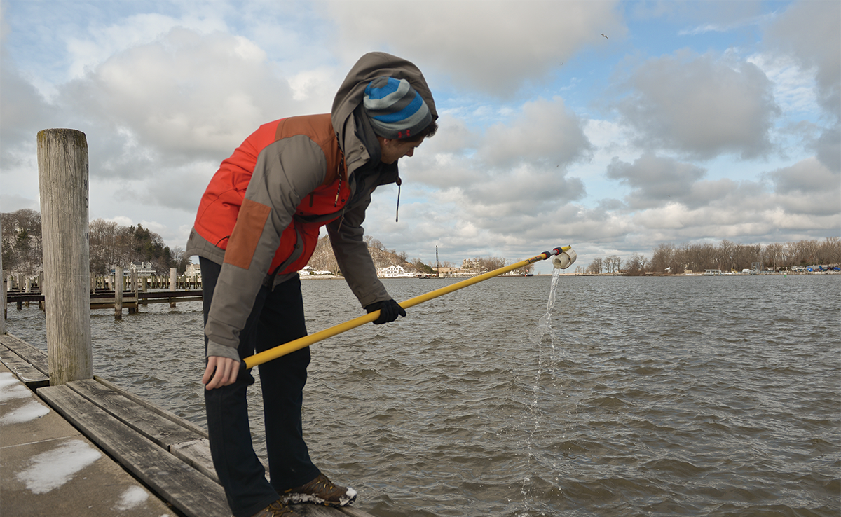 A Hope College student standing on a pier in the winter takes a water sample from Lake Macatawa. He holds a receptacle at the end of a long yellow pole to dip into the water.