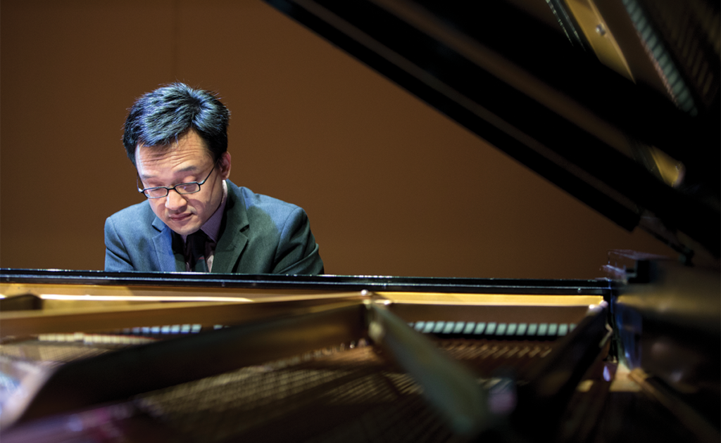 Dr. Andrew Le plays piano. Wearing glasses and a blazer, we see Dr. Lee from the opposite end of a grand piano, through the open lid, beyond the strings.