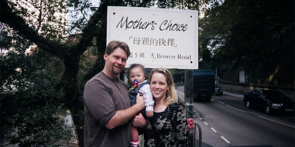 Dr. Dennis Feaster pictured with his wife Sarah, as they adopt their son from his native Hong Kong. They stand with him as a newborn, in front of the Mother's Choice sign which is in both English and Chinese.