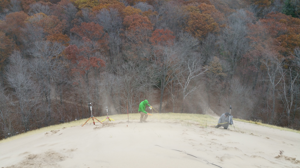 Two geologists set up ultrasonic anemometers on the side of a very windy beach dune. Sand swirls around them. Leafless and fall-color trees are in the background.