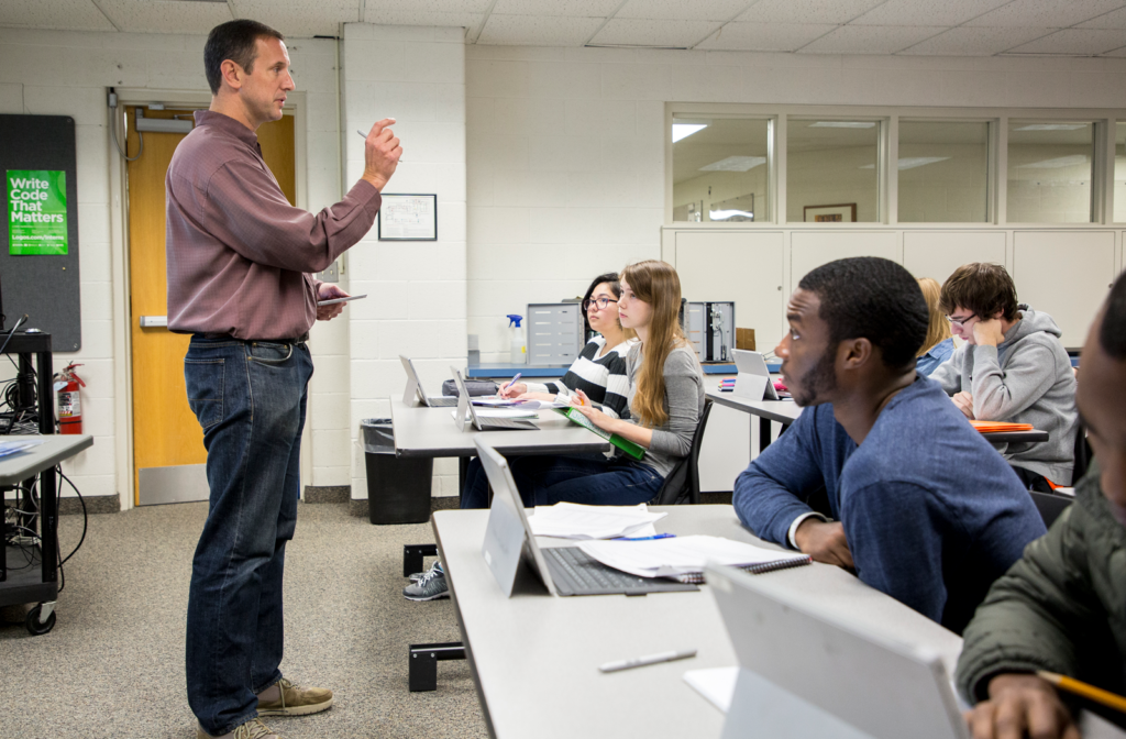 Dr. Ryan McFall stands at front of computer science classroom. Students with laptops on the table in front of them look on attentively, while taking notes. A green poster in the background reads 'Write Code that Matters'.